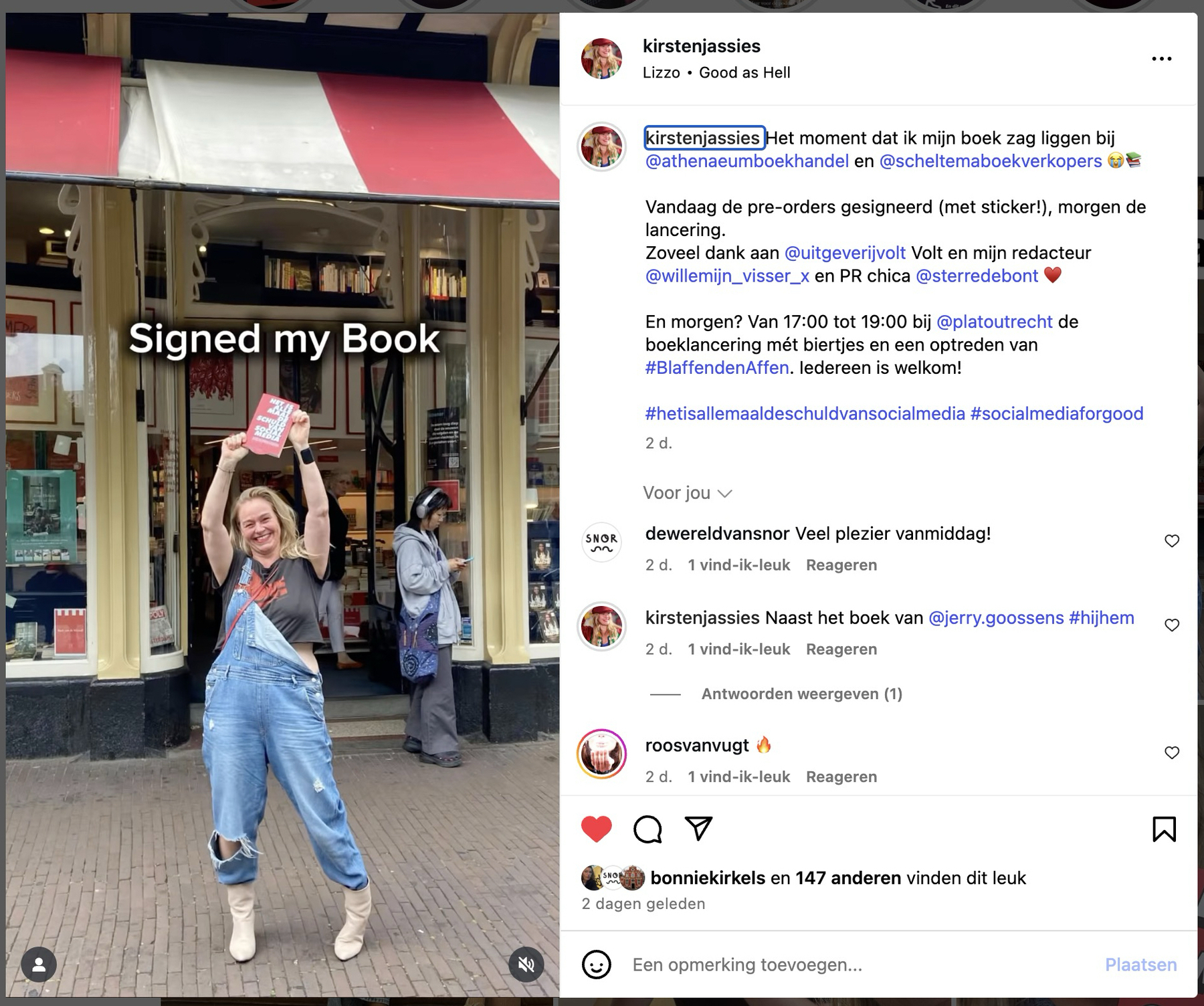 Kirsten poseert vrolijk buiten een boekwinkel terwijl ze een boek omhooghoudt met de tekst ‘Signed my Book’ omringd door een drukke straat en mensen in de achtergrond.
