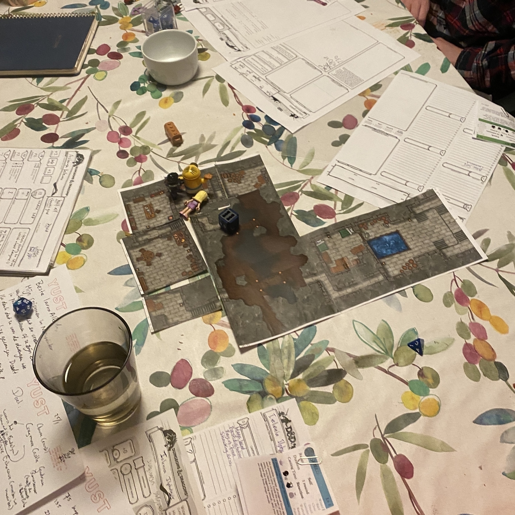 Een bordspel ligt op een tafel met kleurrijke bloemen terwijl spelers miniaturen en kaarten gebruiken om het spel te spelen naast een glas met de drank.