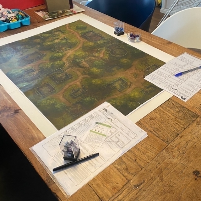 A tabletop setup features a game board with a detailed map and various game pieces, accompanied by notes and a pen on the table.